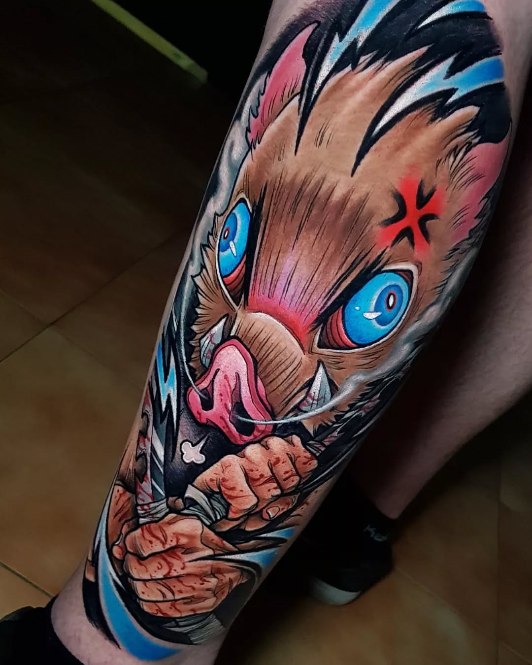 Colorful tattoo on a forearm featuring a brown and pink creature with blue eyes, black spikes, red markings, and clawed hands against a blue background.