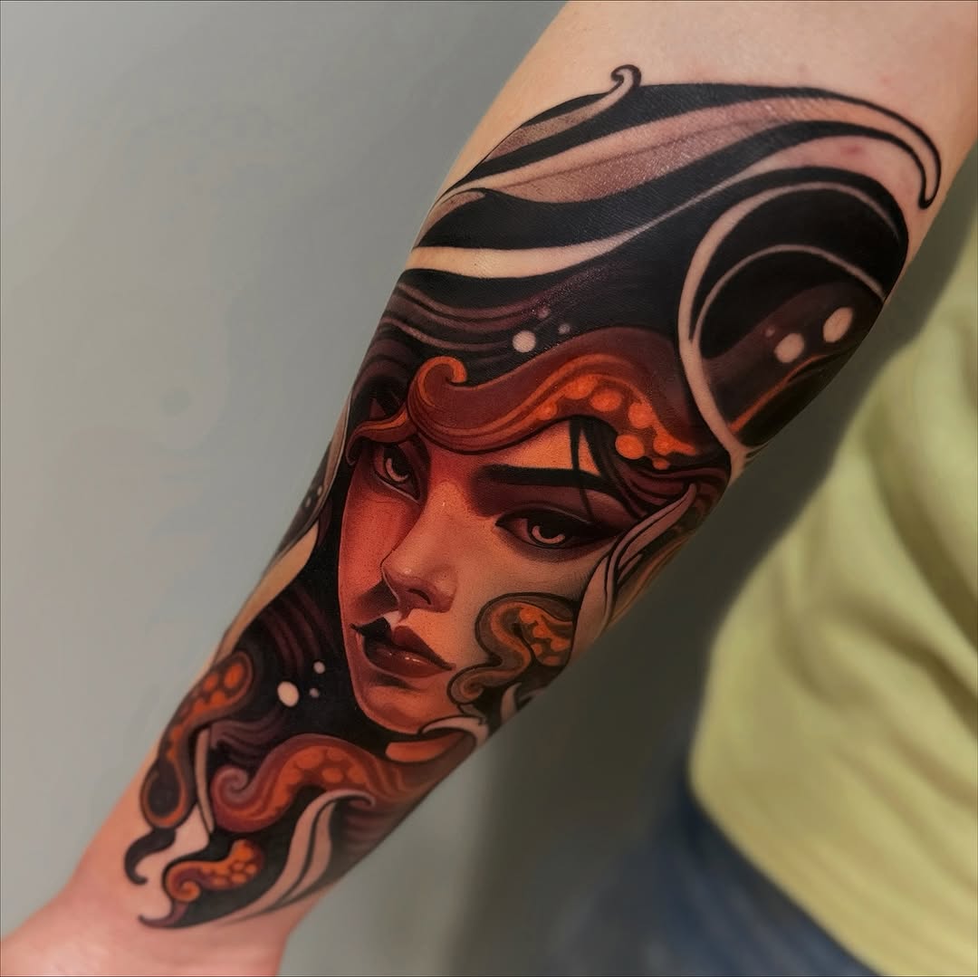 A colorful portrait tattoo on the forearm featuring a face with red and orange tones, ornate swirling details, and dramatic eye makeup in black and dark burgundy.