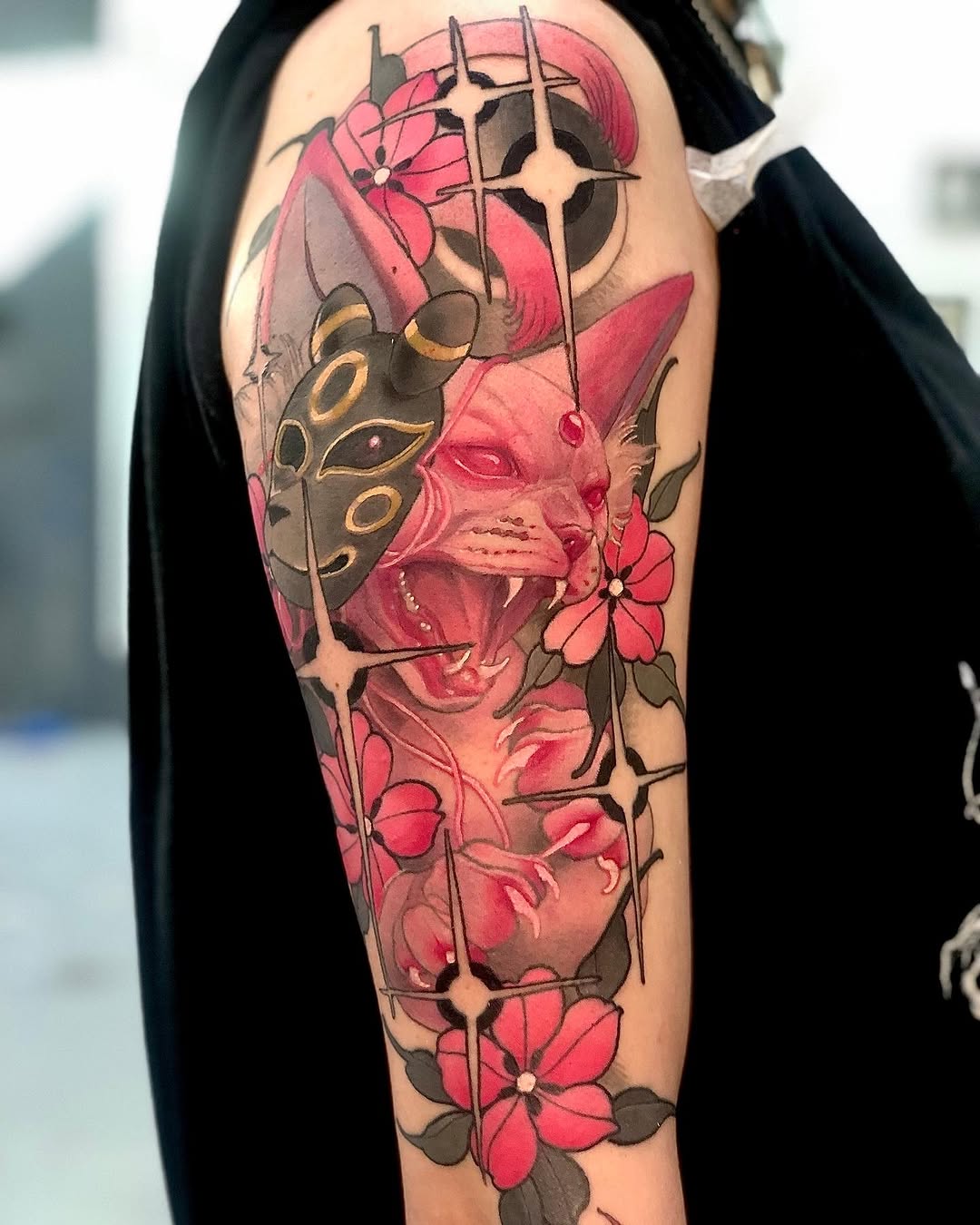 Colorful upper arm tattoo featuring a demonic mask with golden accents, pink flowers, and geometric cross designs on exposed skin.