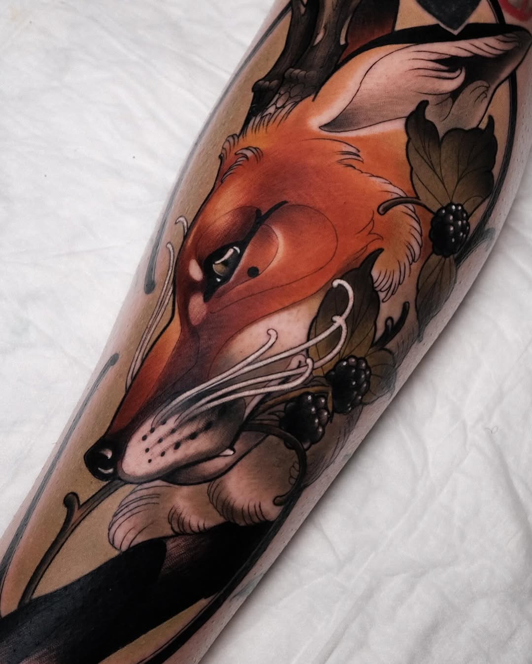 A vibrant red fox head with berries and botanical elements tattooed on an arm, rendered in orange, brown, and black colors.