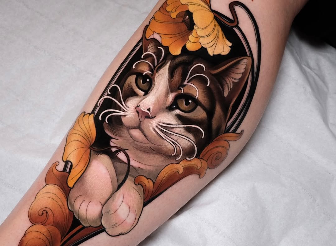 Cat head with brown and tan coloring, large eyes, and white whiskers, framed by ornate orange and black scrollwork, tattooed on an arm.