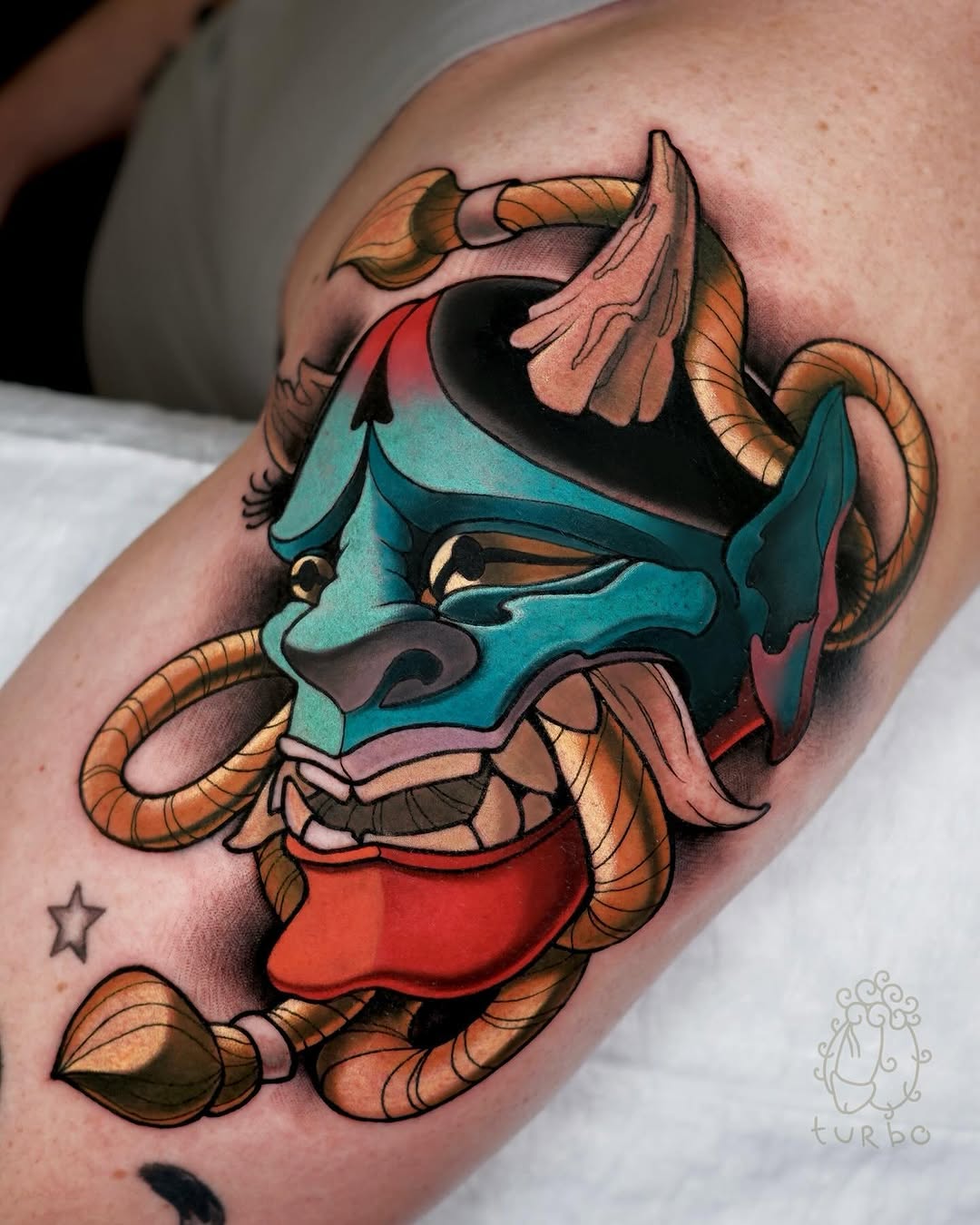 Colorful demon or oni mask tattoo on upper arm featuring turquoise, orange, red, and gold hues with curved horns and expressive facial features.