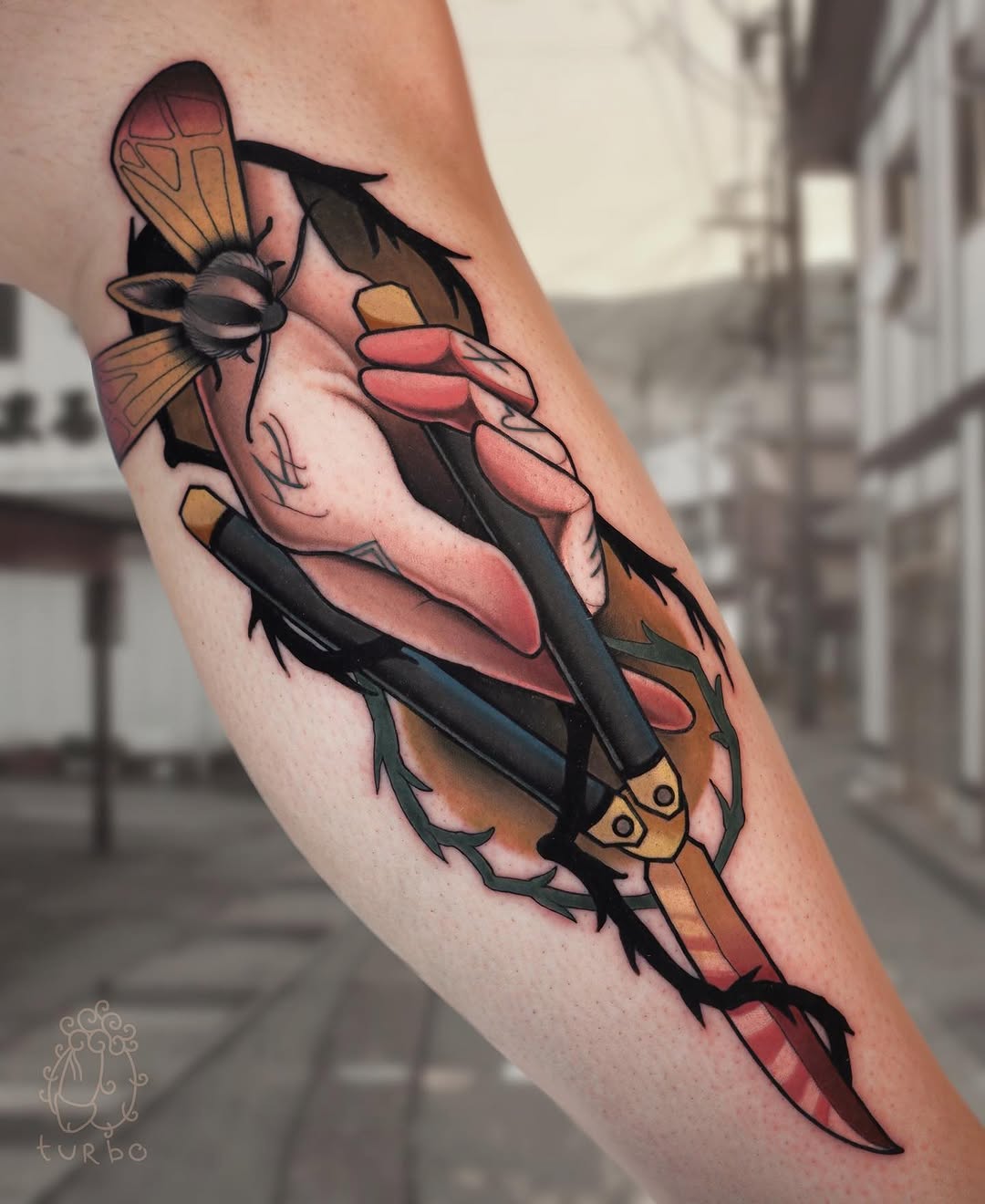 Colorful tattoo on forearm featuring two insects with wings and a hand holding a dark object, in coral, gold, and black tones.