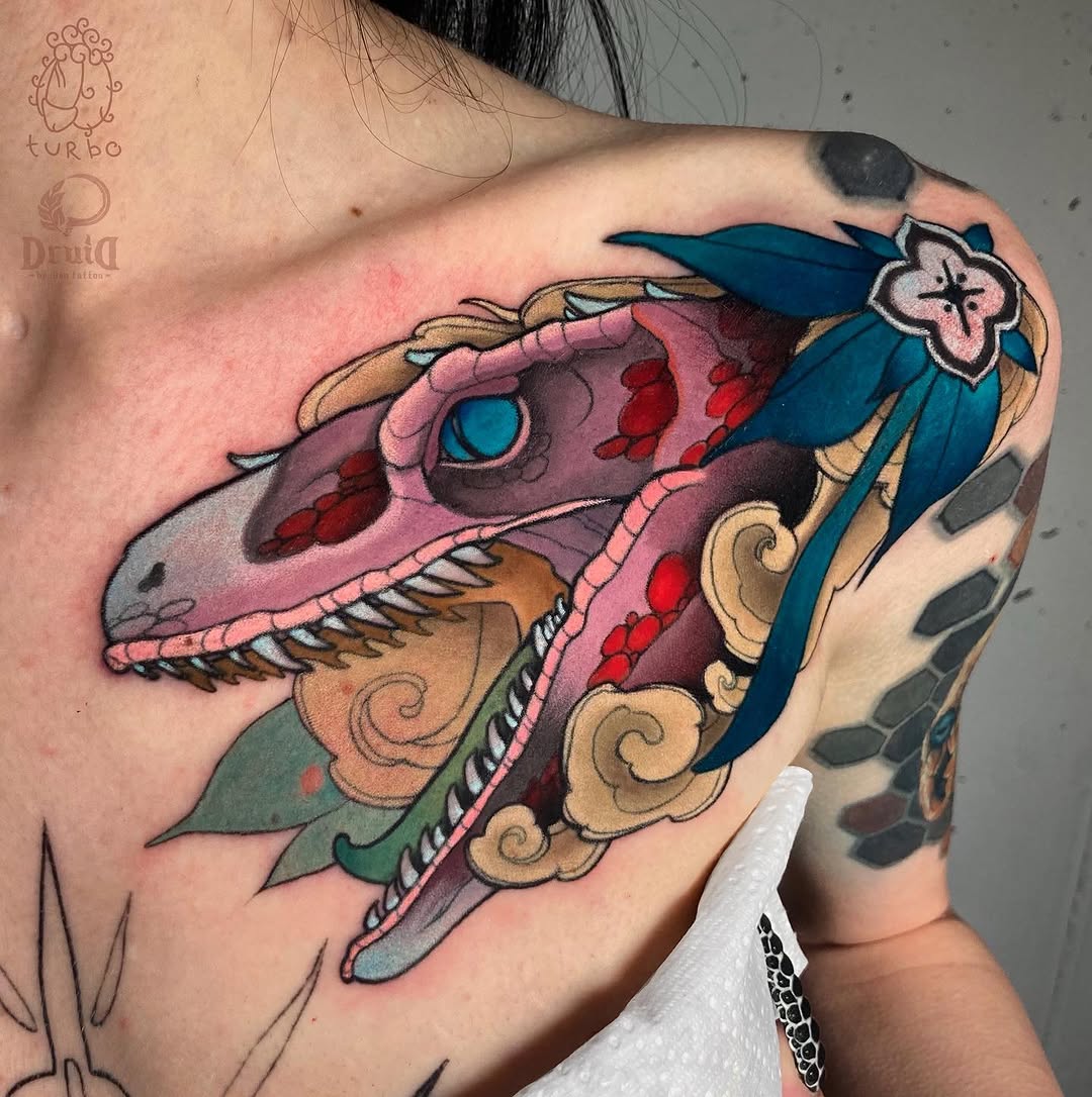 Colorful dragon head tattoo on shoulder featuring pink, teal, and gold ornamental details with red spots and decorative scrollwork.
