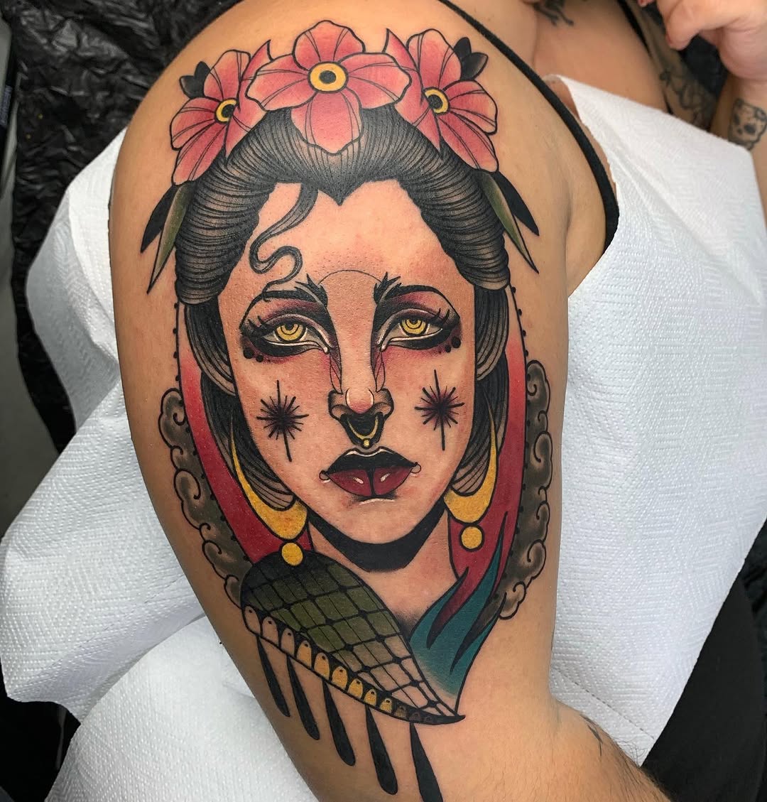 Colorful shoulder tattoo featuring a woman's face with yellow eyes, red lips, star cheek markings, flower crown, gold earrings, and winged collar detail.
