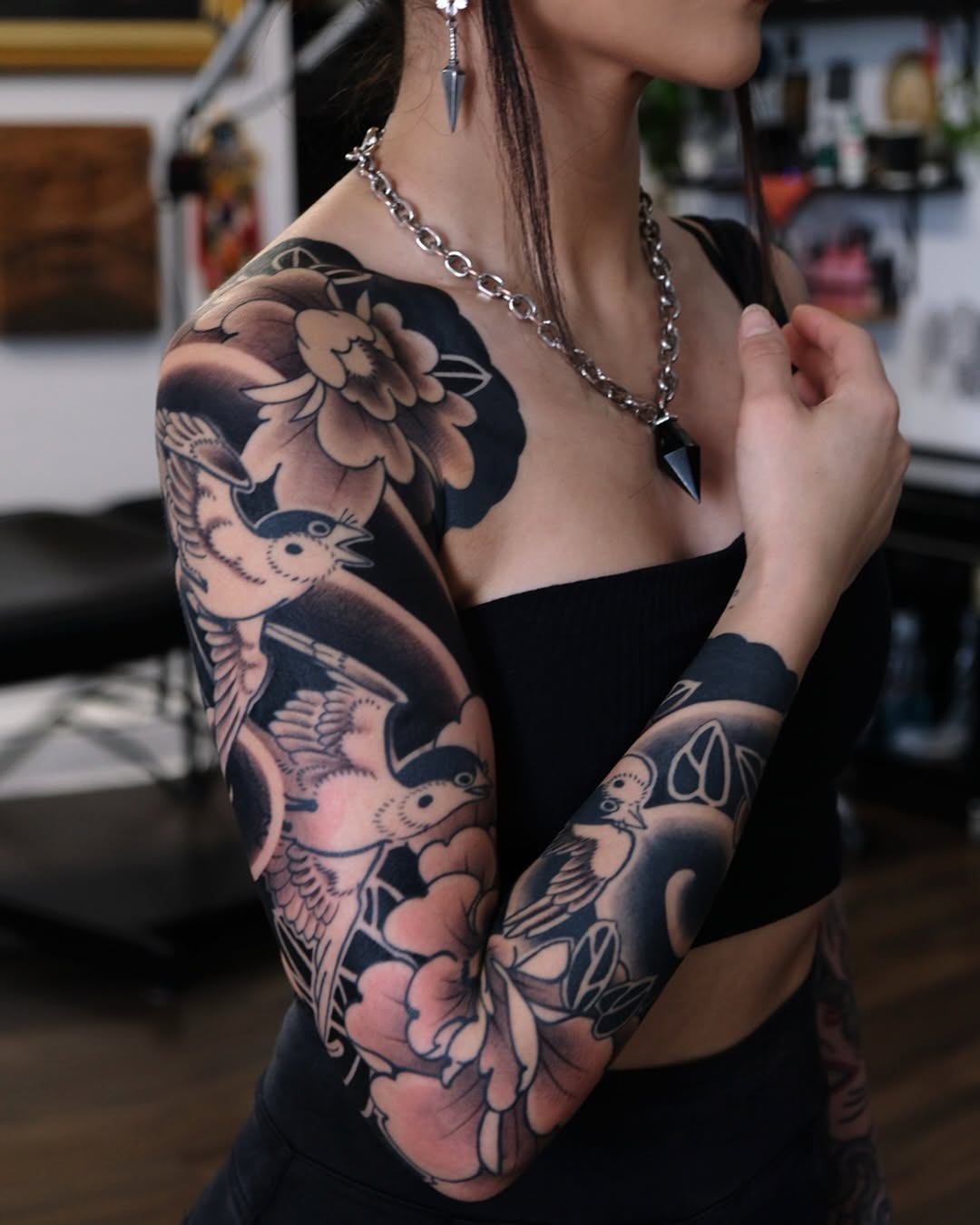 Woman displaying a full arm sleeve tattoo featuring birds, flowers, and botanical elements in black ink with shading on her shoulder and forearm.