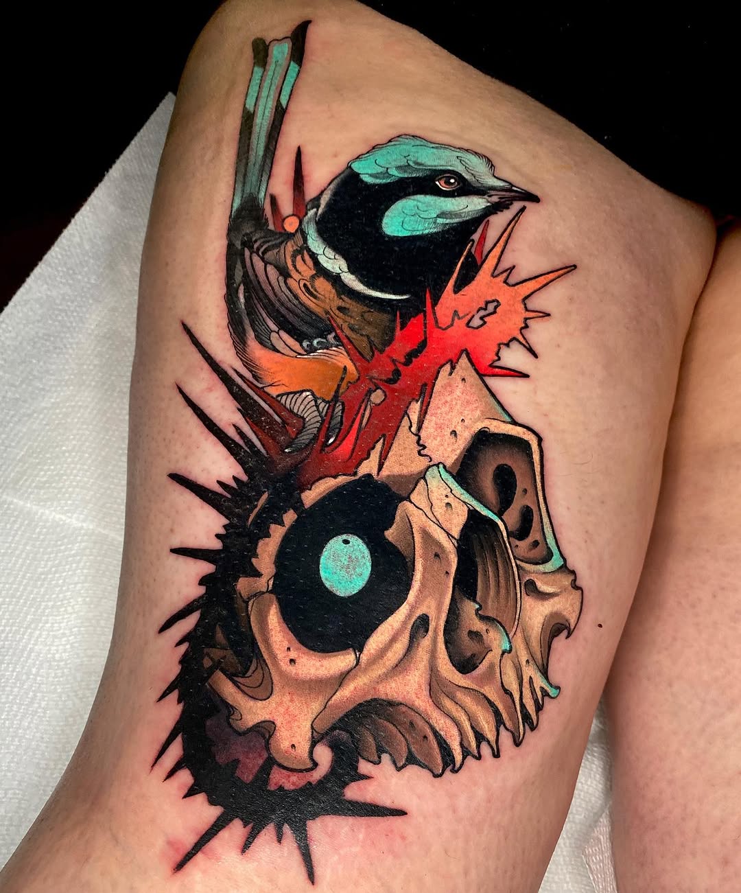 Colorful bird with turquoise head perched on a skull with red spikes and black spines, tattooed on upper thigh.
