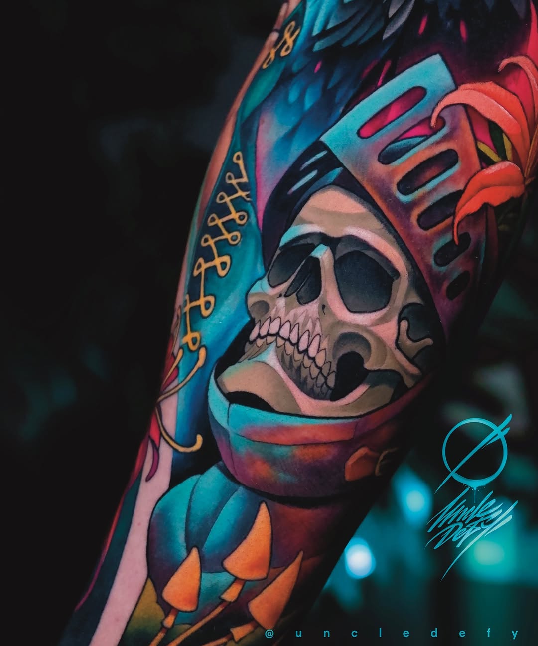 Colorful skull tattoo on arm wearing ornate turquoise and red headpiece with gold patterns, vibrant blues, oranges, and purples throughout.