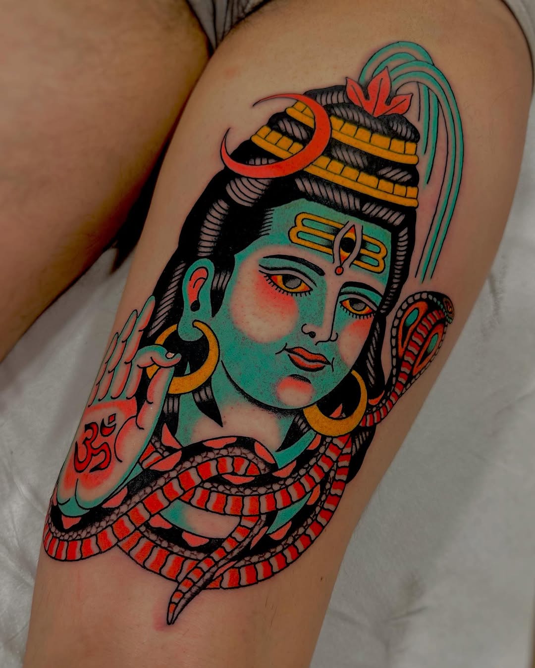 Colorful tattoo on upper leg featuring a deity figure with turquoise skin, red lips, gold jewelry, and a serpent wrapped around the body in red and black.