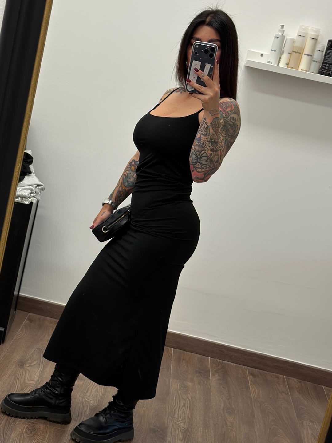 Woman displaying large colorful floral and decorative tattoos covering both arms, photographed in mirror selfie wearing black dress.