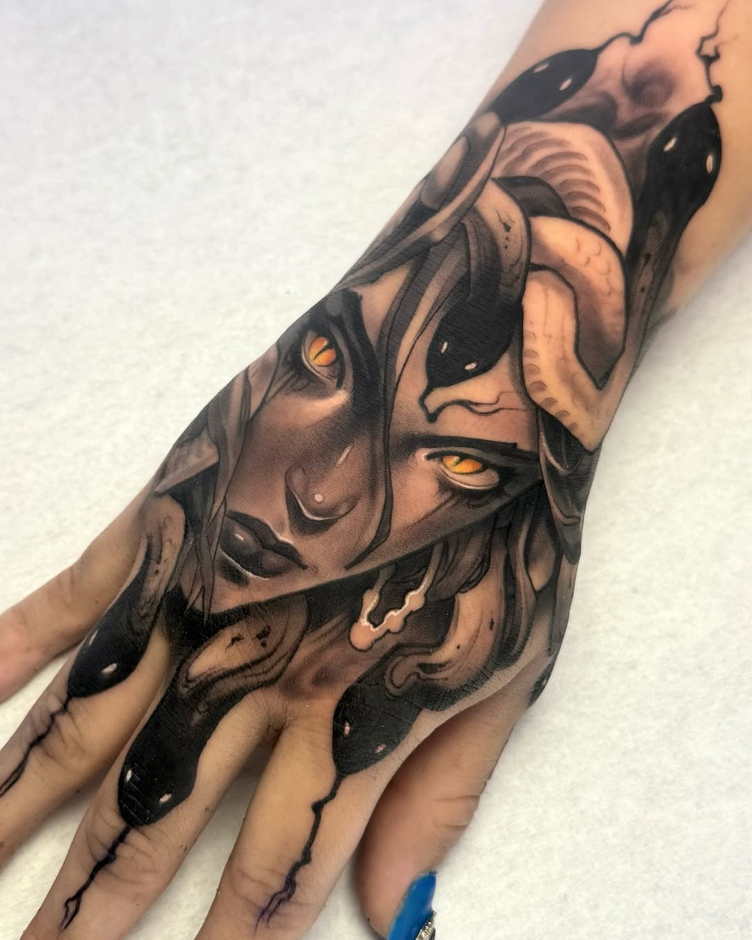 Detailed portrait tattoo on the hand featuring a woman's face with striking yellow eyes, dark hair, and skull imagery in black and gray ink.