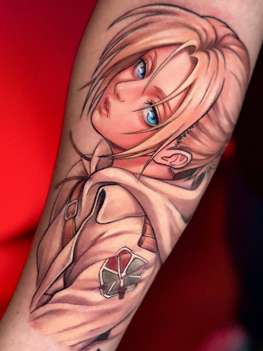 Colored arm tattoo featuring a character with peach-toned hair and blue eyes, with a smaller emblem below on the bicep against a red background.