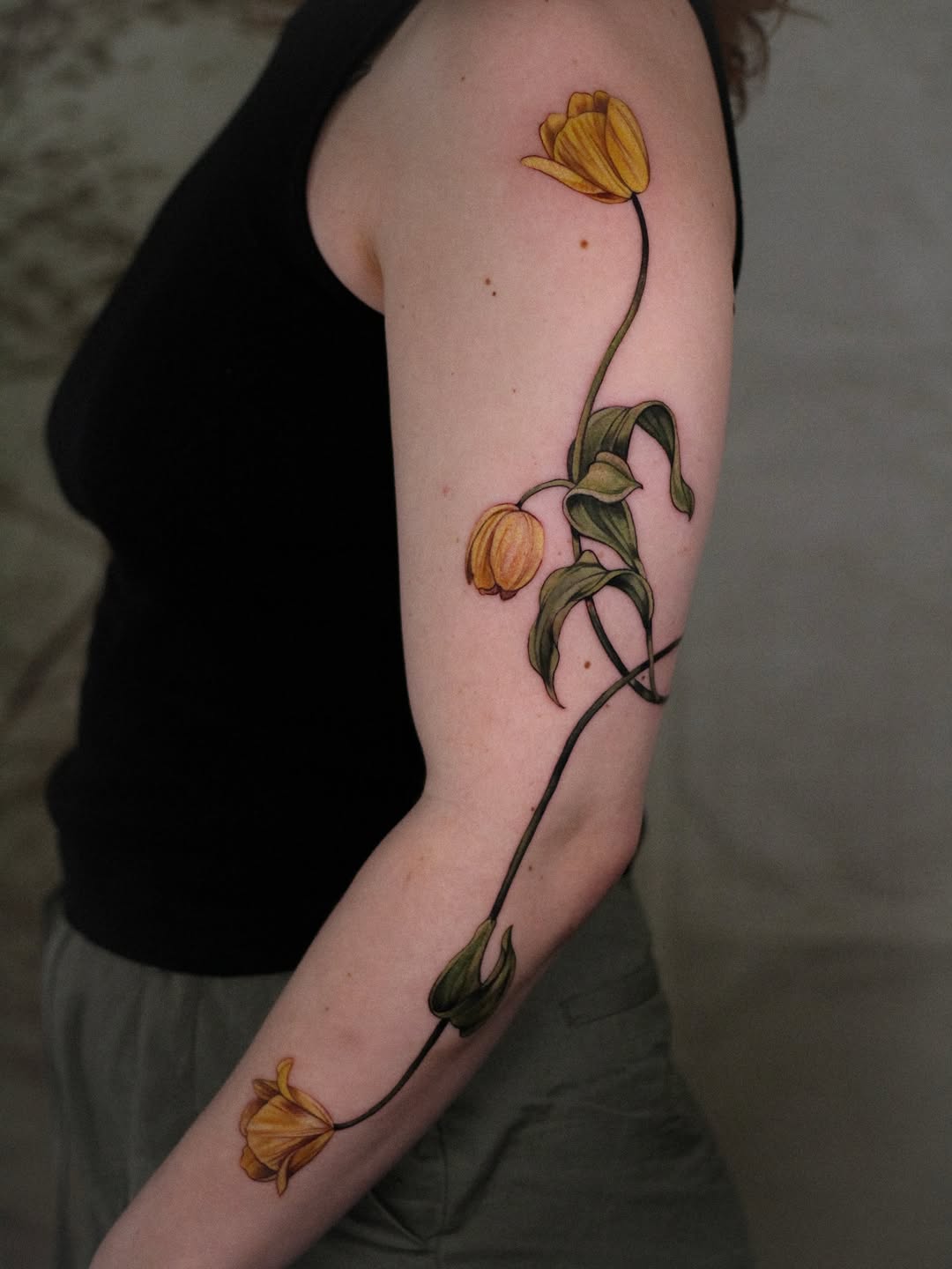 Vertical arm tattoo featuring yellow tulips in various stages of bloom connected by a curved green stem with leaves.