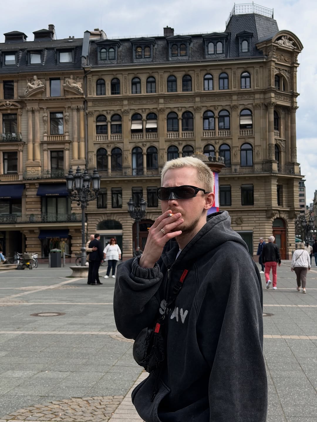 A person wearing dark sunglasses and a black hoodie stands in an urban square with a classical building behind them.