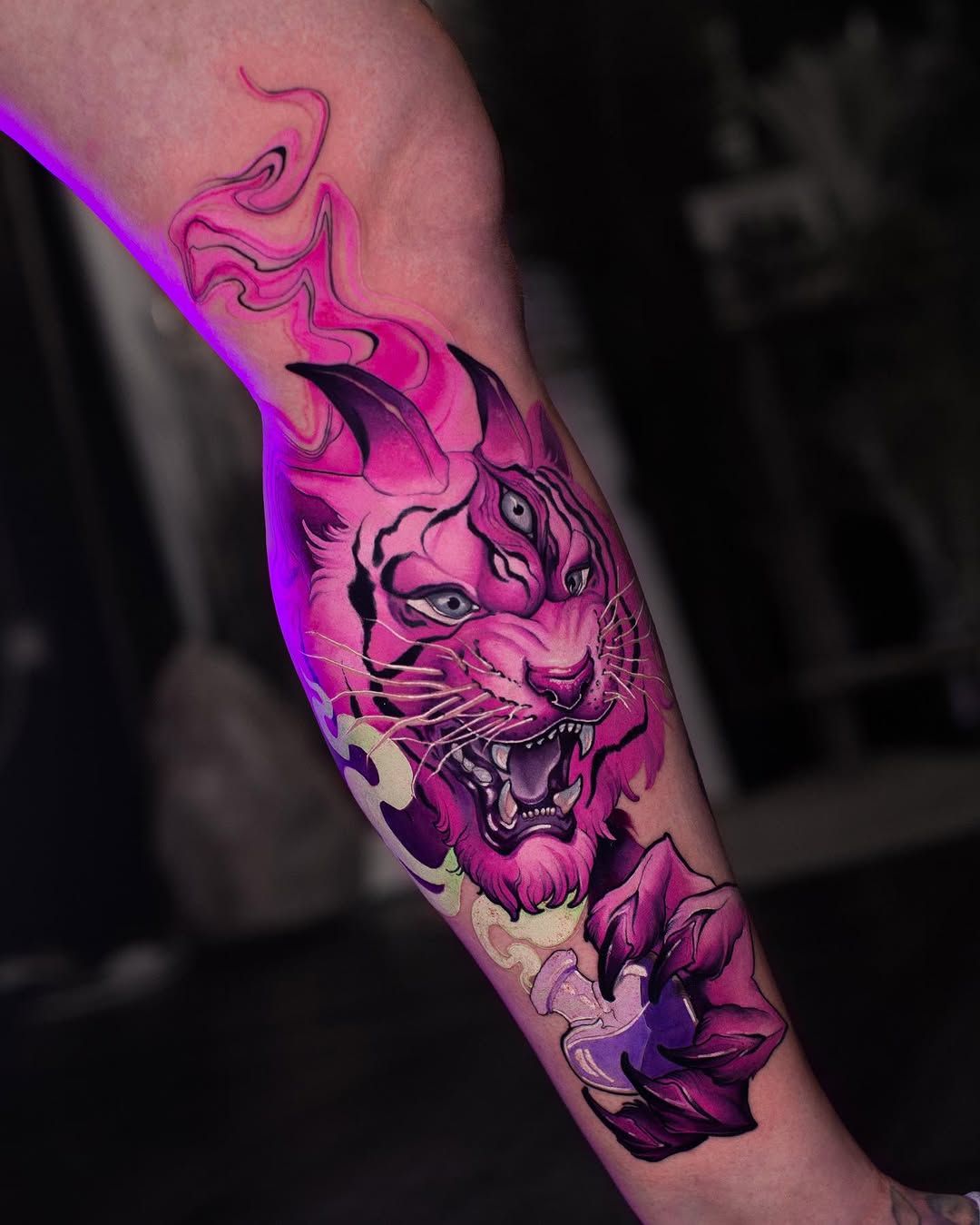 A vibrant pink and purple lion head tattoo with flowing flames above it and floral elements below, positioned on the inner forearm.