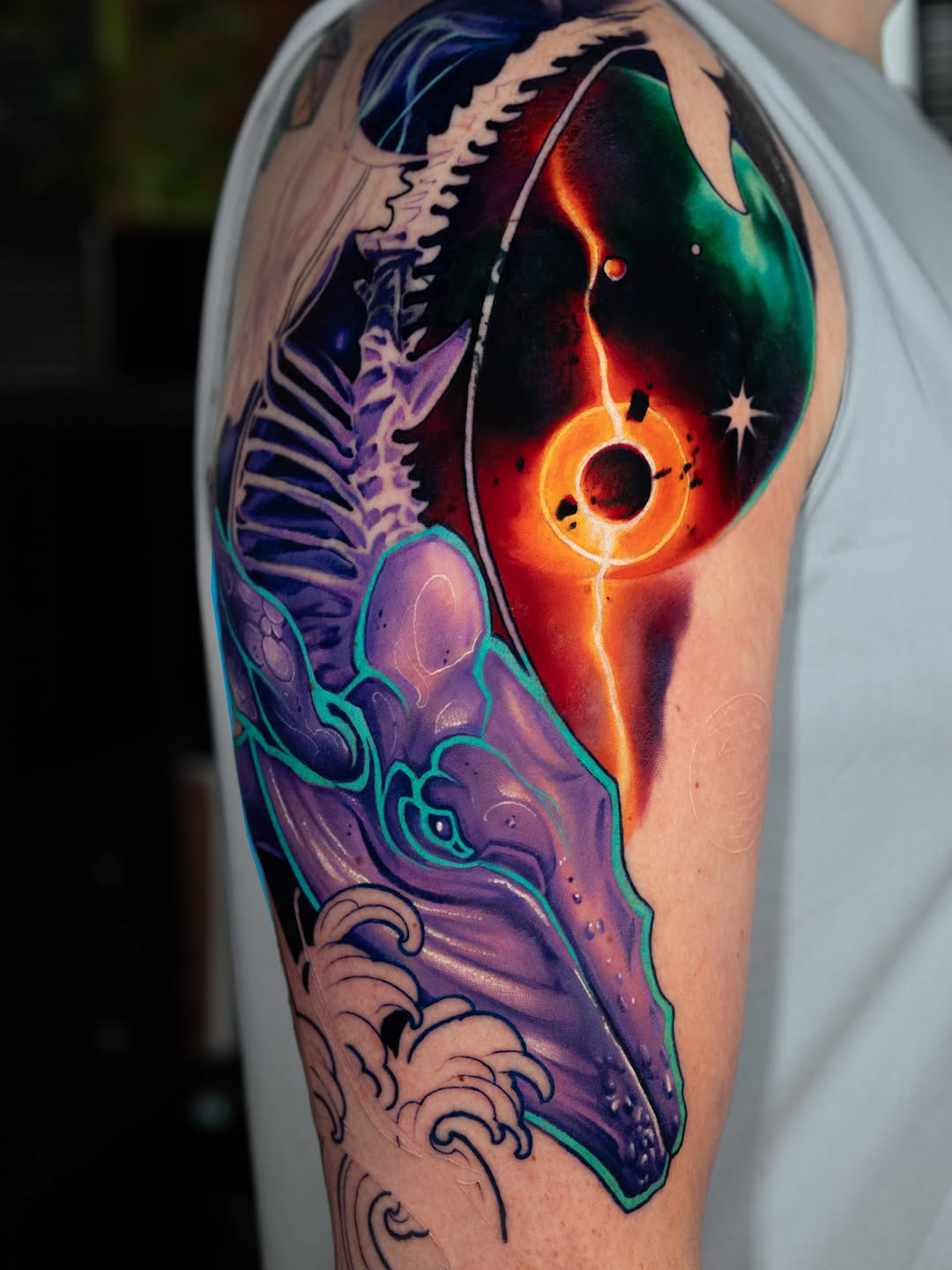 Colorful shoulder tattoo featuring a glowing sun with planetary rings, skeletal ribcage, purple anatomical organs, clouds, and ornamental swirls in vibrant oranges, purples, greens, and blues.