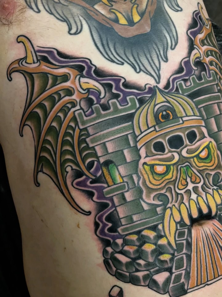 Colorful skull tattoo with ornate decorative elements, wings, and facial features in purple, green, yellow, and orange on the upper arm and shoulder.