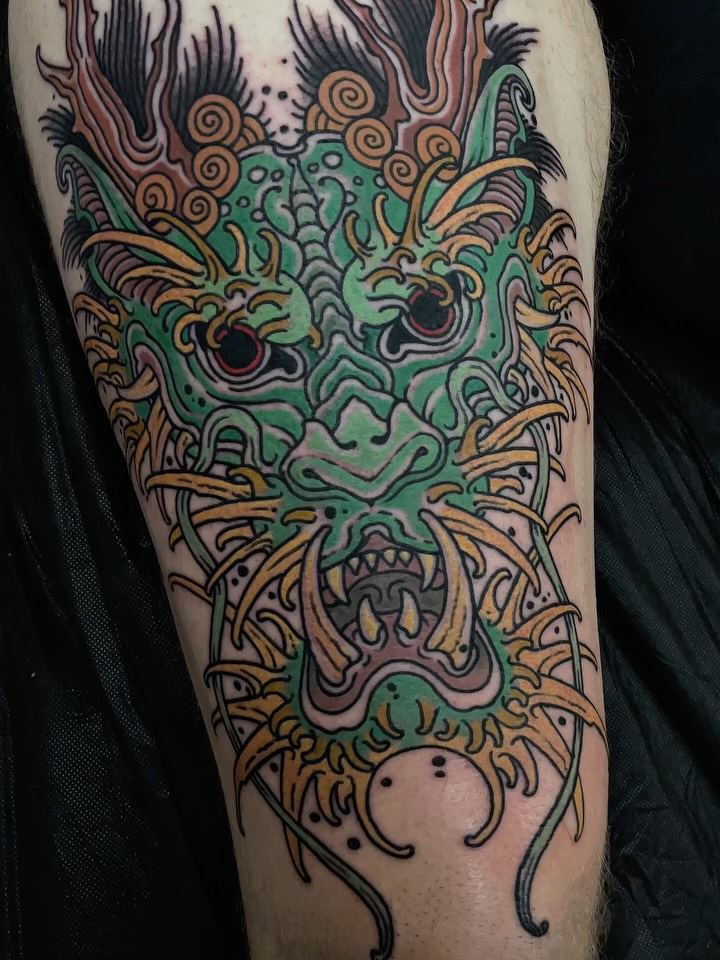 Colorful demon or oni mask tattoo on thigh featuring green face, orange and black decorative elements, and ornamental details in gold, green, pink, and black ink.