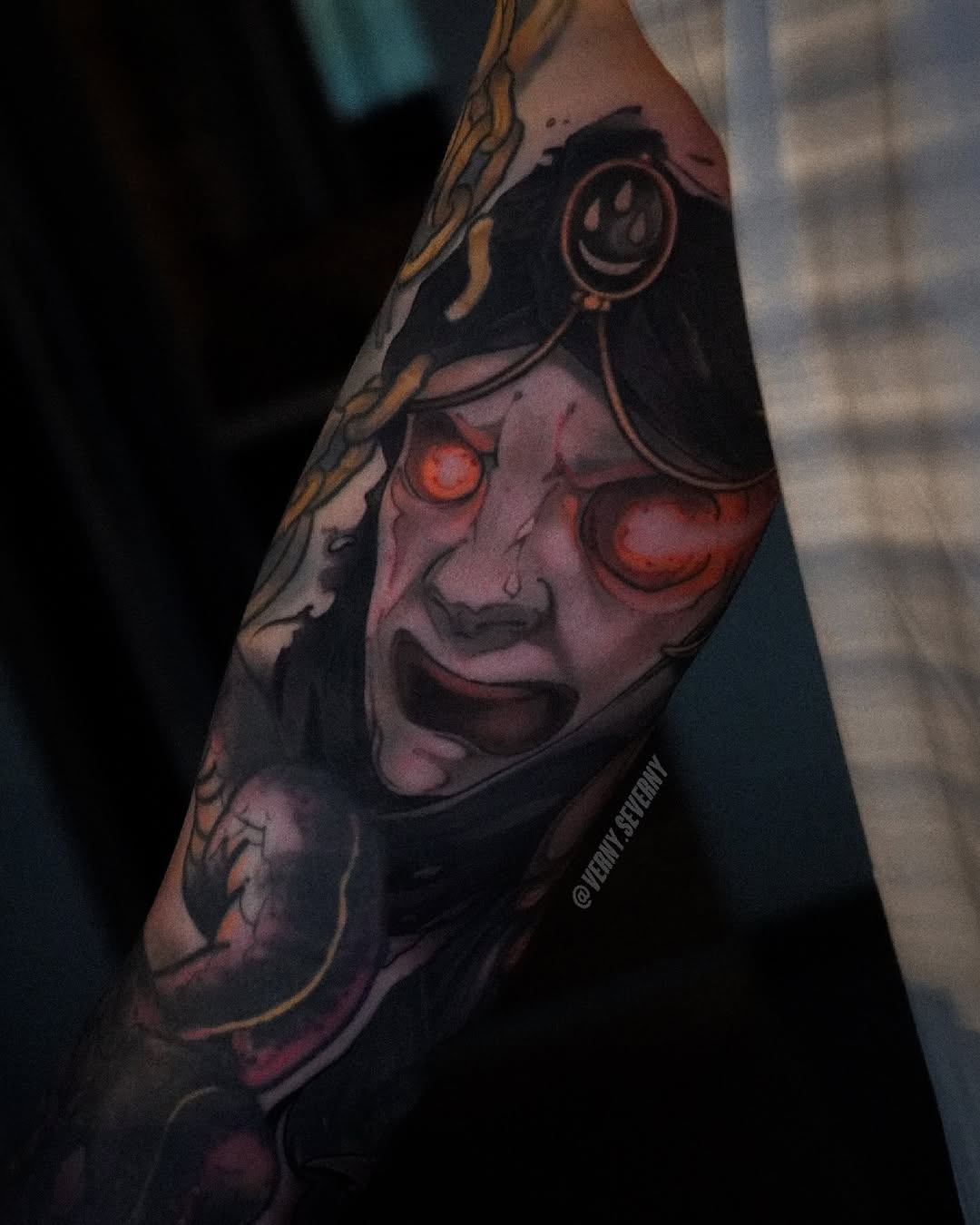 Colored tattoo on forearm featuring a face with glowing red eyes, a smiley face pendant, black hood, and gold decorative elements.
