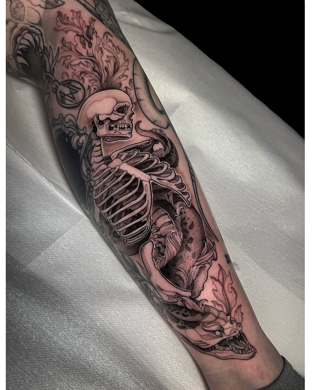 Skeletal anatomy tattoo on forearm featuring a skull and ribcage with ornamental scrollwork flourishes in grayscale and tan tones.