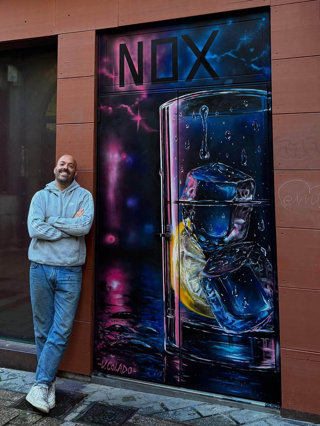 Man with crossed arms standing beside storefront door displaying neon NIX logo and ice cube artwork in pink, blue, and yellow colors.