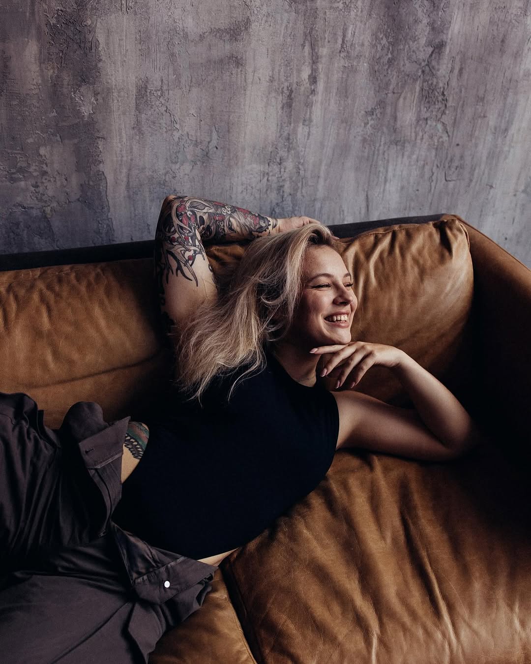 Woman with a large colored arm tattoo featuring organic flowing designs in red and black tones, seated on a tan leather couch.