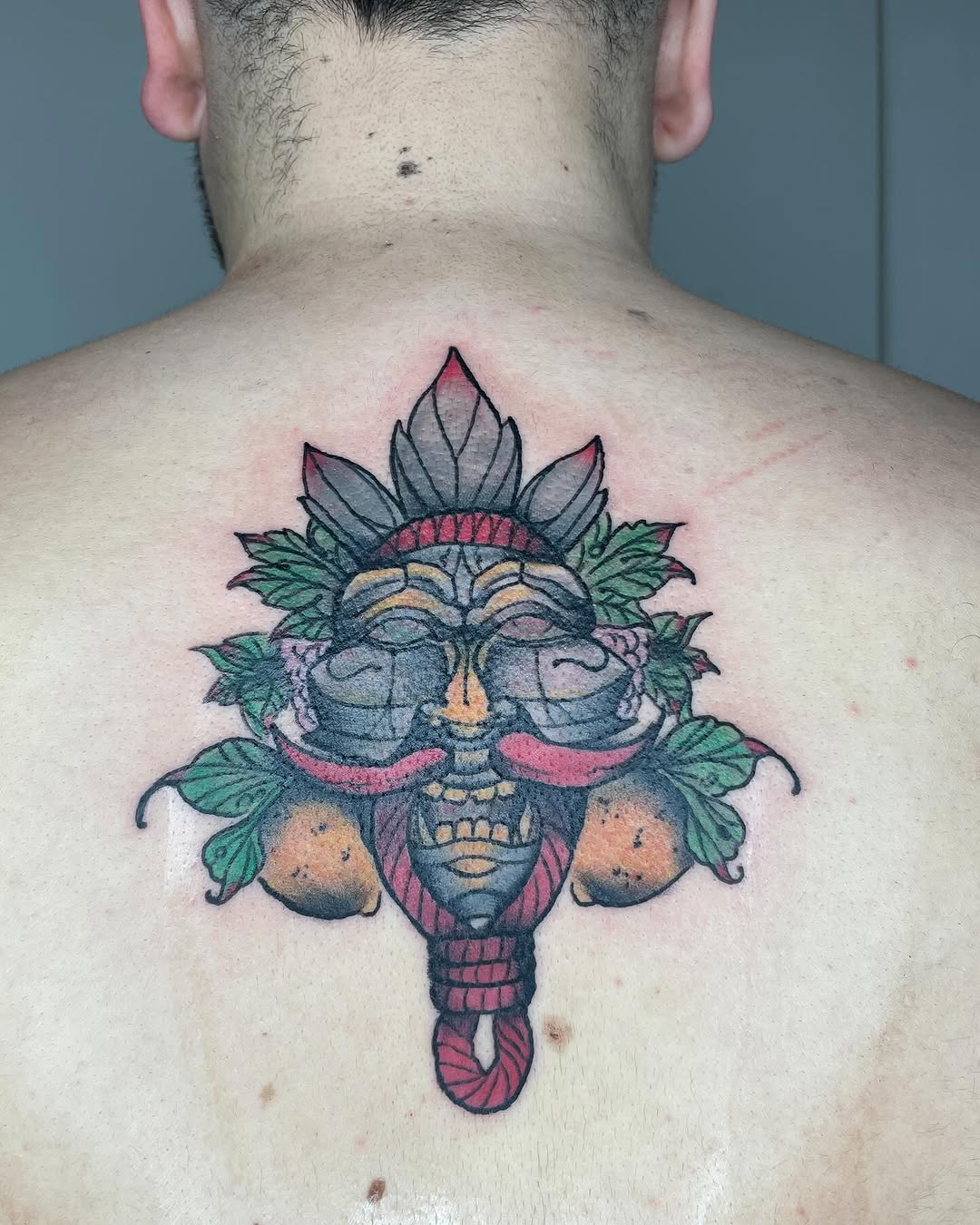 Colorful mask-like face tattoo with feathered headdress, green leaves, and dangling ornaments on upper back in red, blue, green, and gold tones.