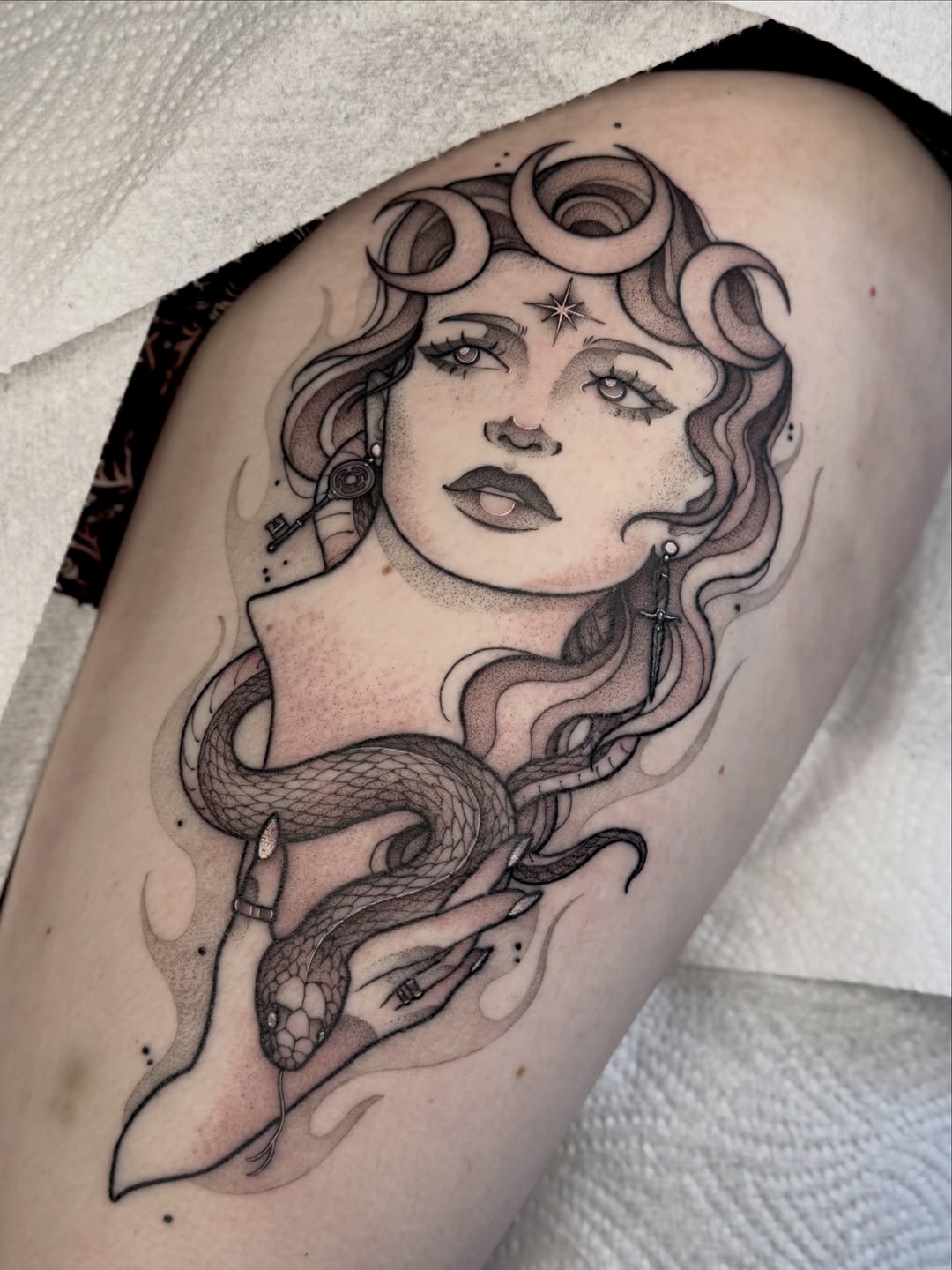 Woman's face with wavy hair and closed eyes, intertwined with a coiled snake below, rendered in black ink on thigh.