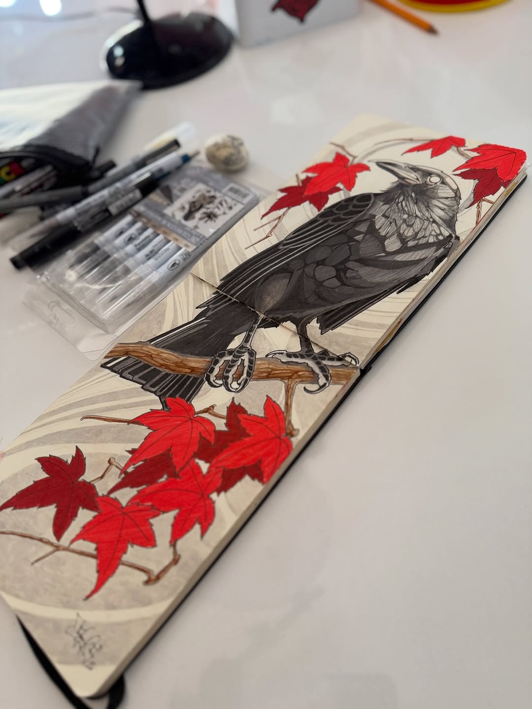 Black crow perched on a brown branch with red maple leaves above and below, displayed in an open sketchbook surrounded by art supplies.