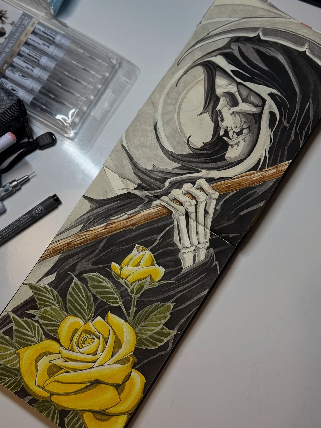 Tattoo design on paper featuring a skull in hooded robes holding a scythe, with yellow roses and green leaves below.