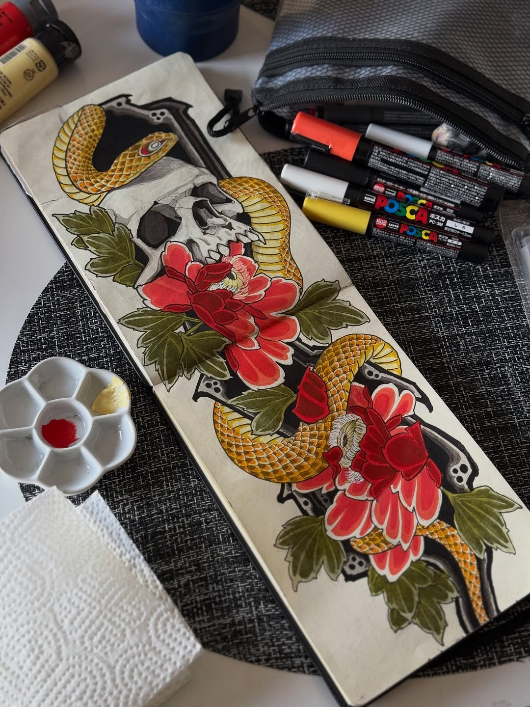 Tattoo design sketch on cream paper featuring two golden snakes intertwined with red peonies and green leaves, surrounded by skull imagery in black ink.