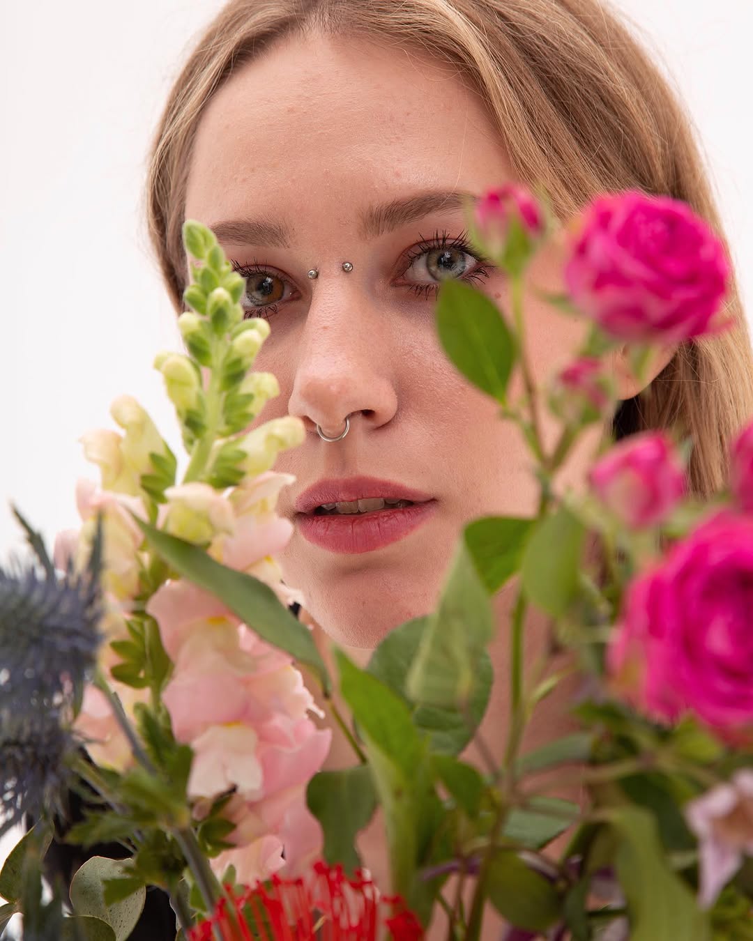 Close-up portrait of a person with a nose ring and small facial piercings, surrounded by pink flowers and green foliage against a white background.