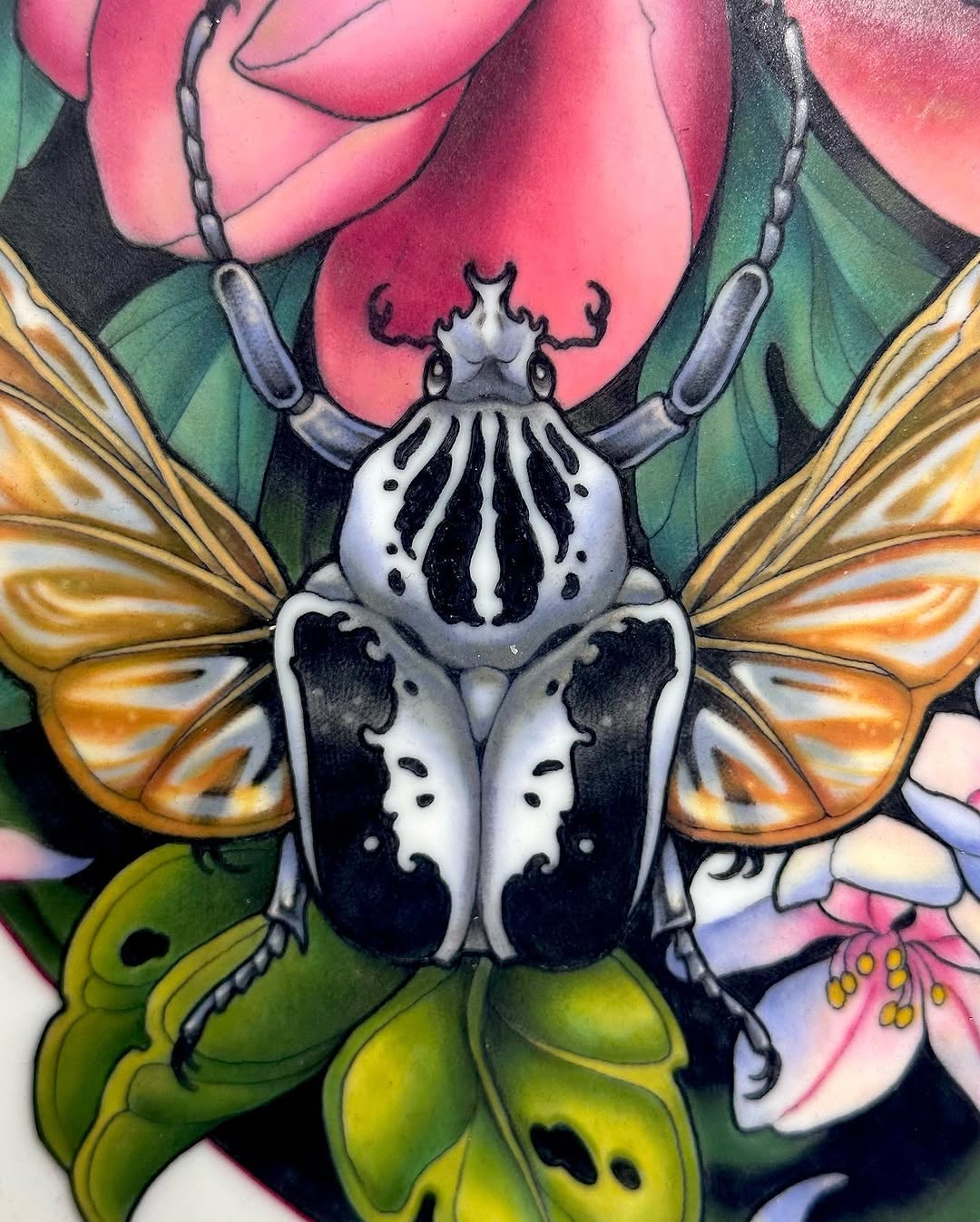 Colorful beetle tattoo with white and black body, golden wings, surrounded by pink roses and green leaves on the upper arm.