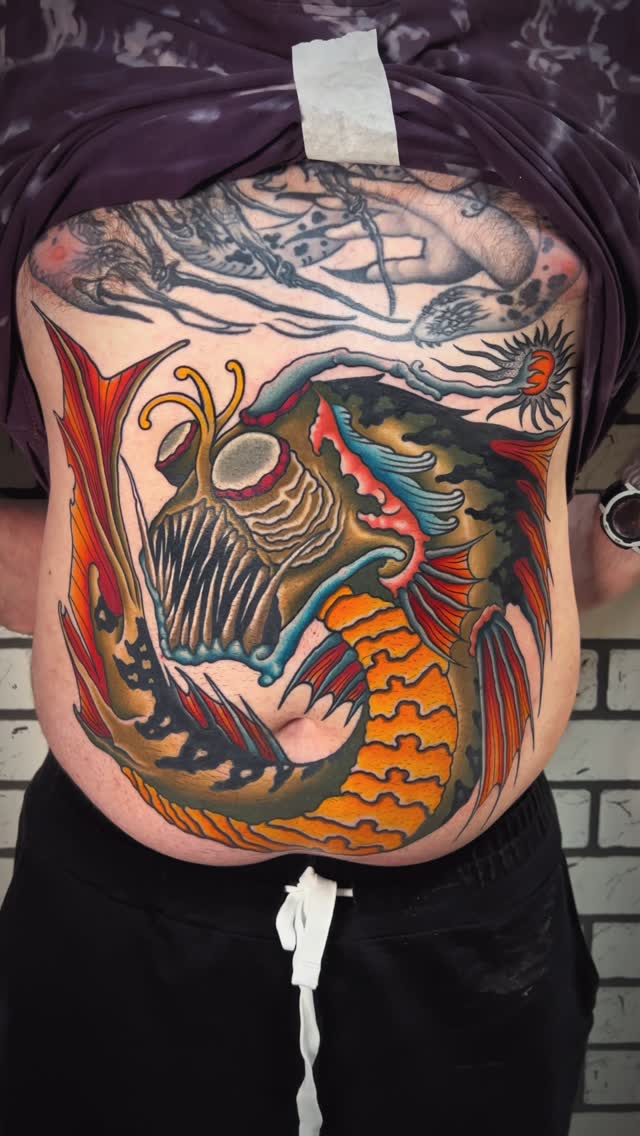 Colorful chest and torso tattoo featuring a mythical dragon-fish hybrid with skull, wings, and ornamental details in red, gold, blue, and black.