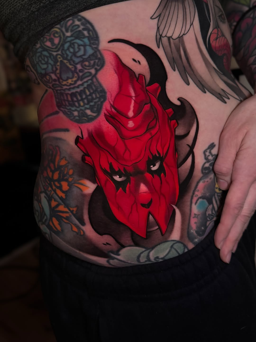 Red demonic face with horns and a woman's profile on upper arm, surrounded by skull, wings, and floral designs in multiple colors.