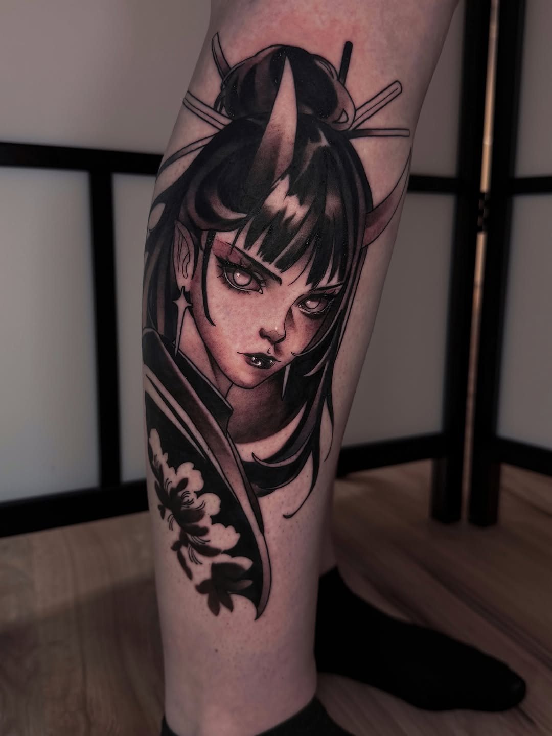 Tattoo on lower leg featuring an androgynous figure with horns, bangs, and facial markings, with a small demon illustration below, rendered in black ink.