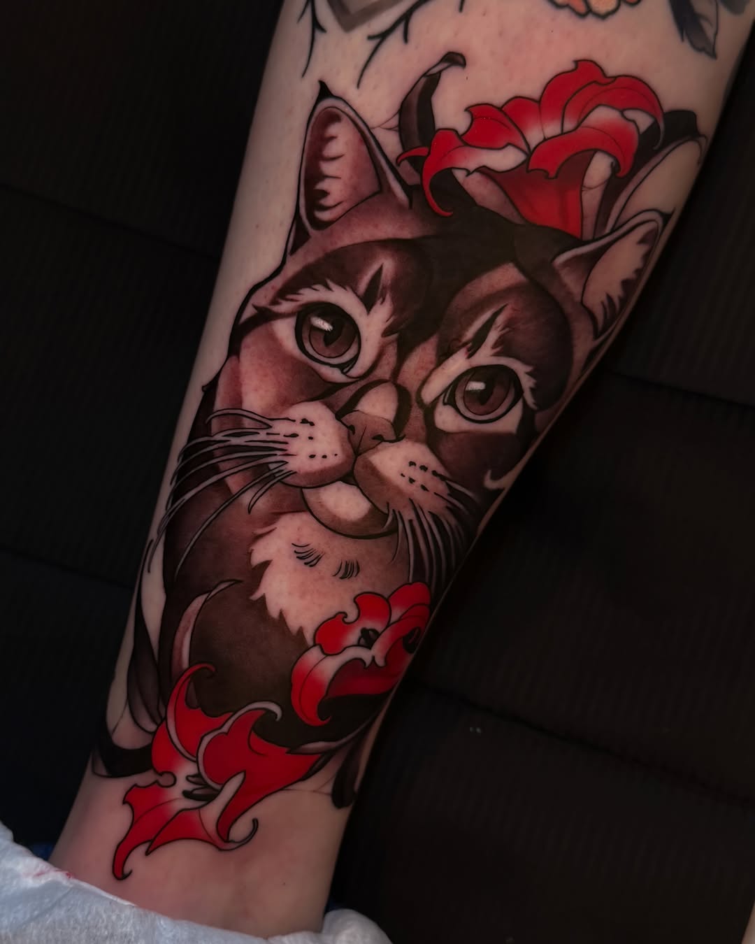 Cat with large eyes and red flowers framing the head and lower body, tattooed on the forearm in brown and red ink.