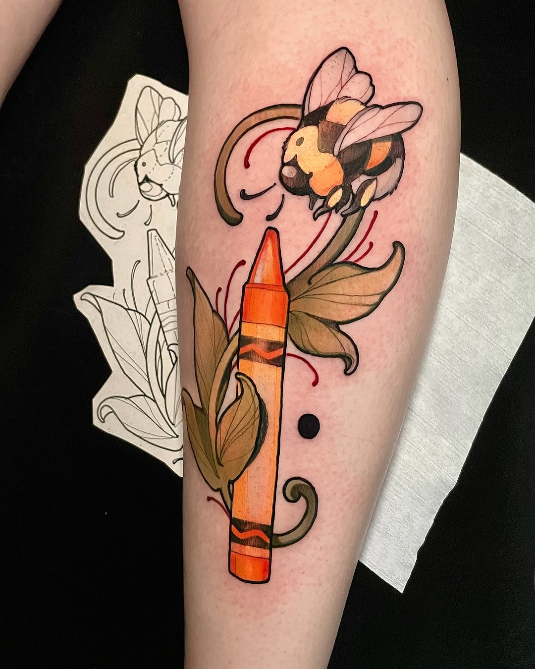 Colorful leg tattoo featuring a bee, bullet with floral elements, and sketch design on white paper held beside it.