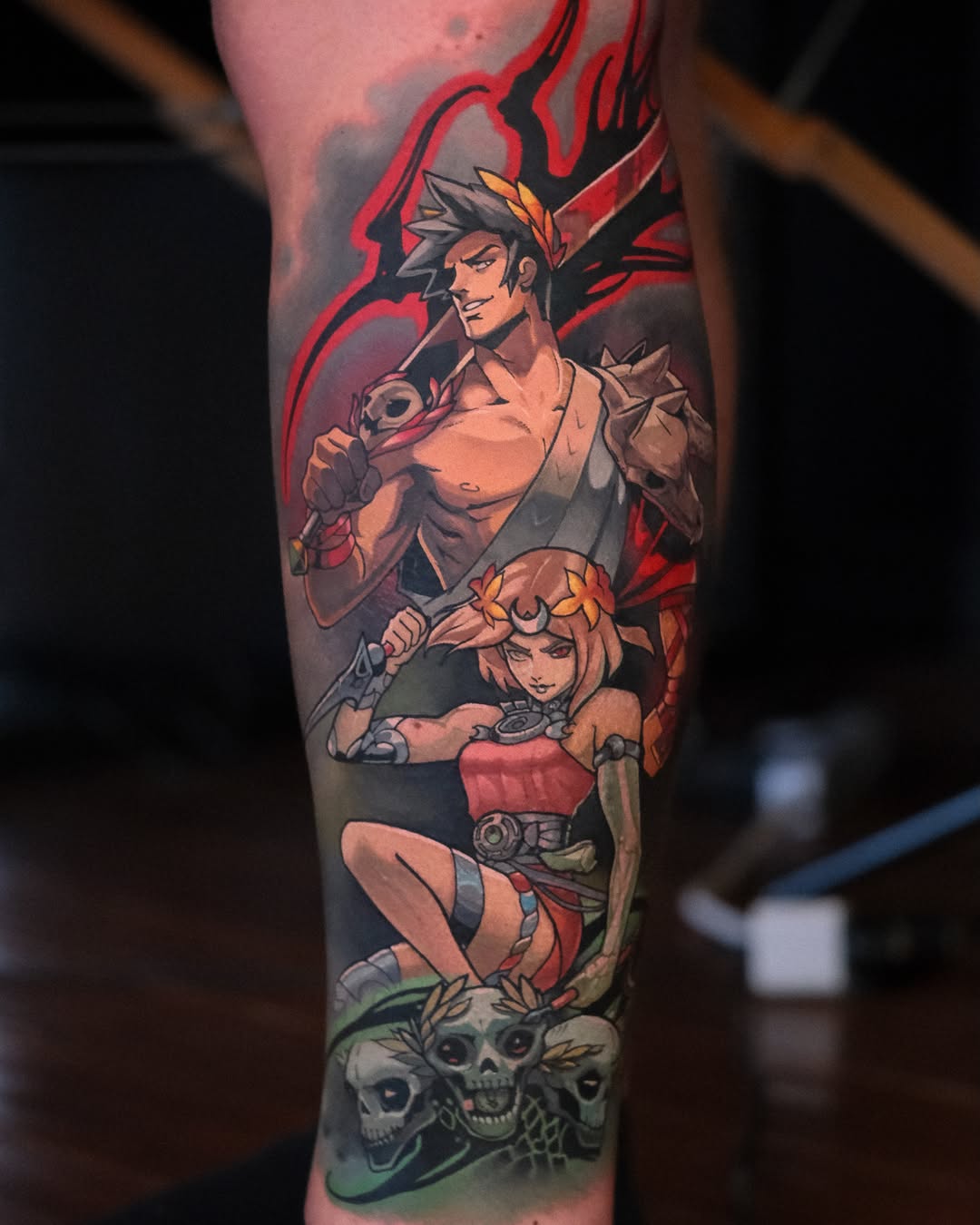 Colorful leg tattoo featuring two animated characters with red and orange accents, skulls at the base, and flame-like designs in the background.