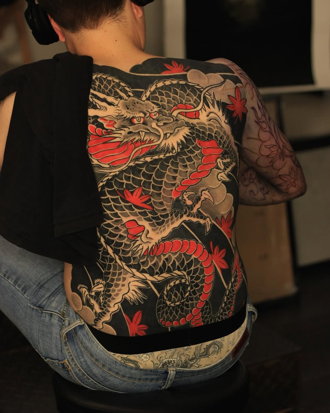 Back piece tattoo featuring two gold and cream dragons with red accents and maple leaves on black background, displayed on shirtless person wearing jeans.