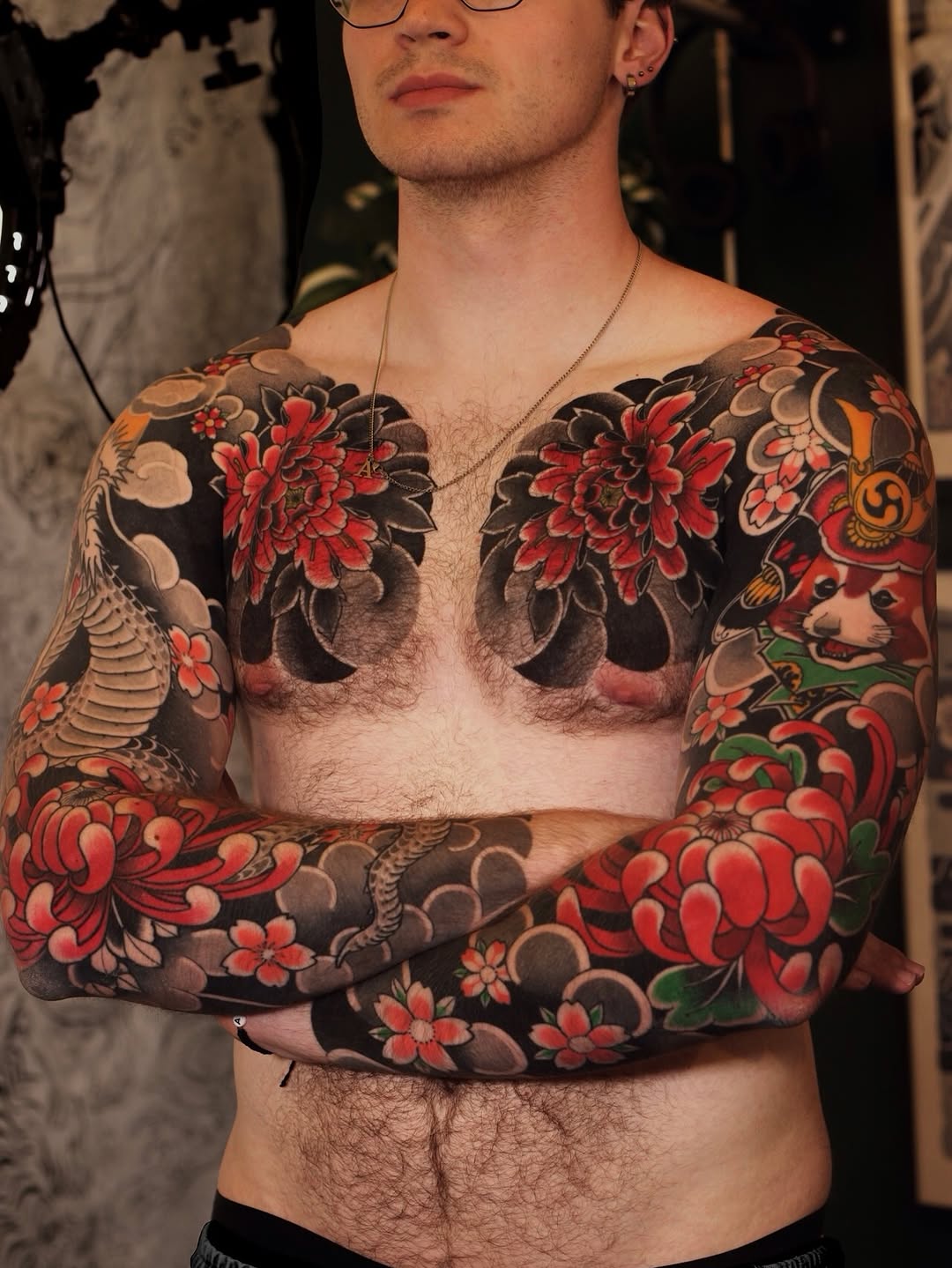Shirtless man displaying full chest and sleeve tattoos featuring red flowers, moons, and botanical elements on dark background in warm tones.