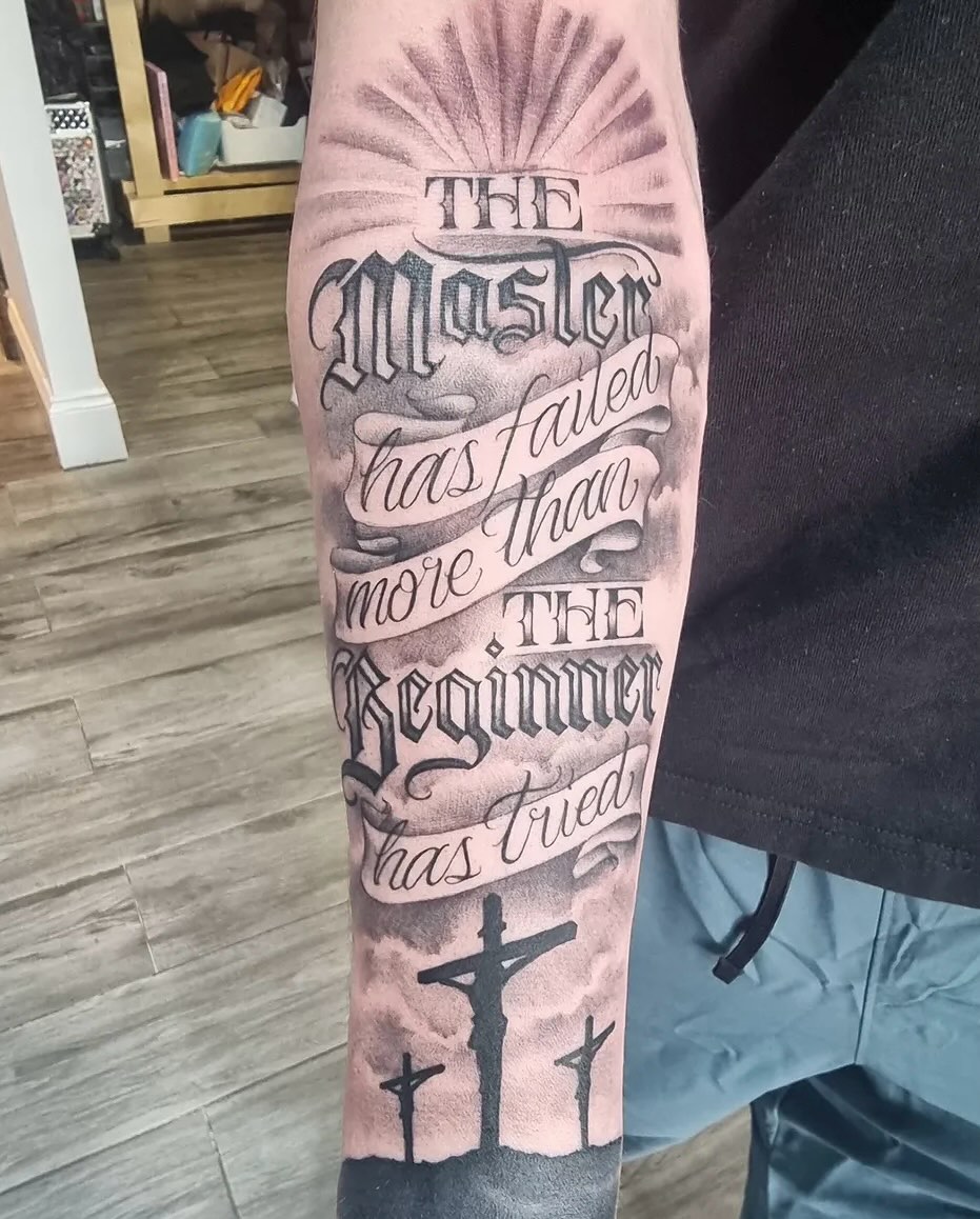 Forearm tattoo featuring religious text with radiating sunburst, script lettering, and three crosses silhouetted against a landscape in gray and black ink.