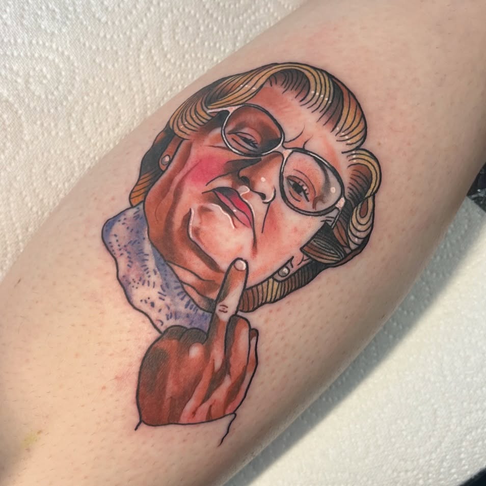 Colored tattoo on arm featuring a person with glasses and styled hair making a peace sign gesture, rendered in red, purple, and brown tones.