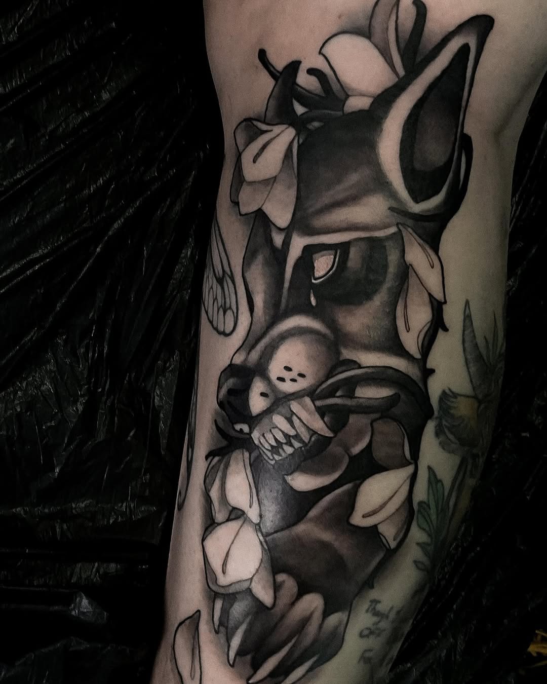Monochrome tattoo on the forearm featuring a rabbit holding a skeletal figure surrounded by flowering plants and botanical elements.