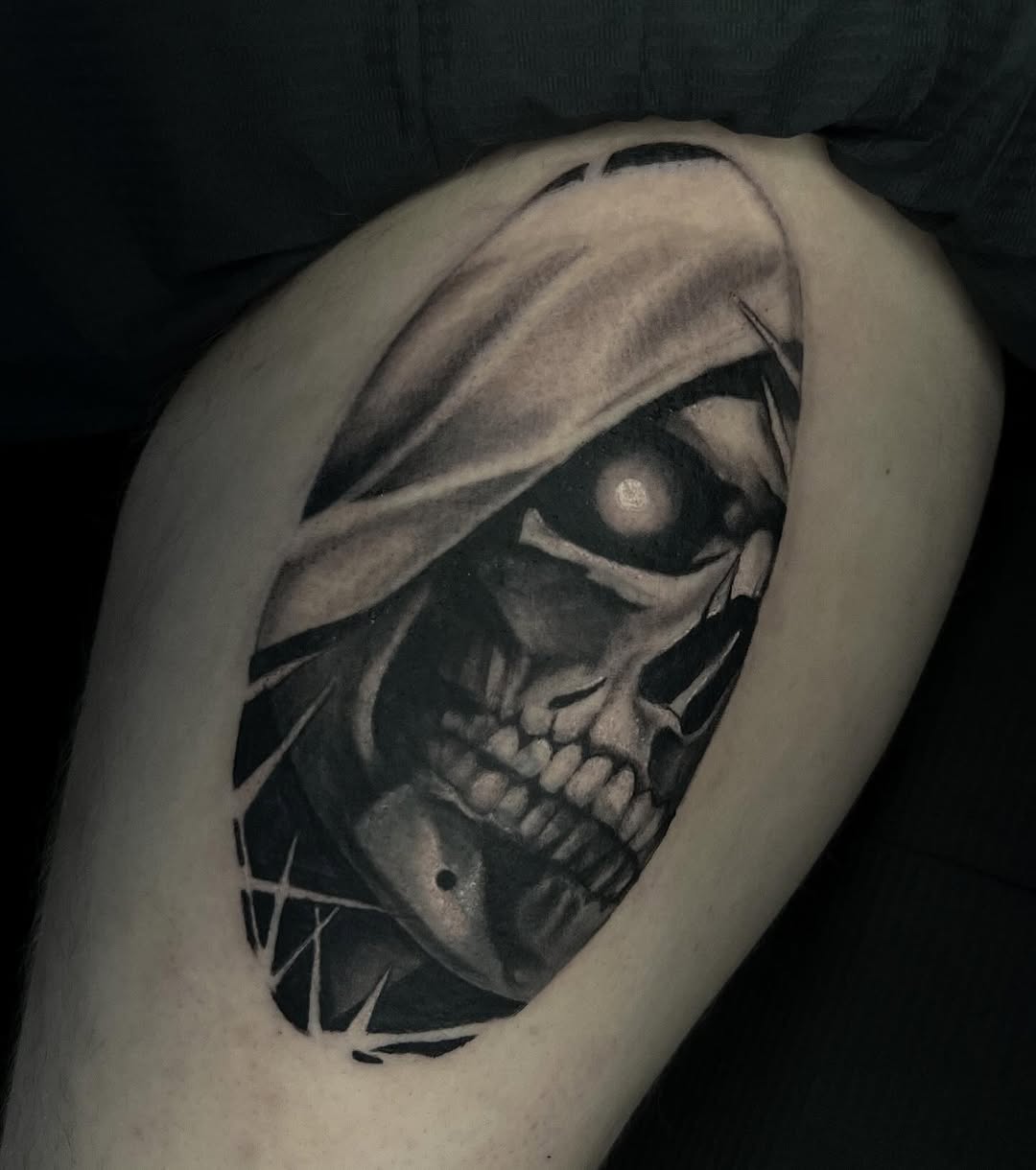 Skull with glowing eye wrapped in flowing fabric on upper arm in black and grey ink with geometric elements.