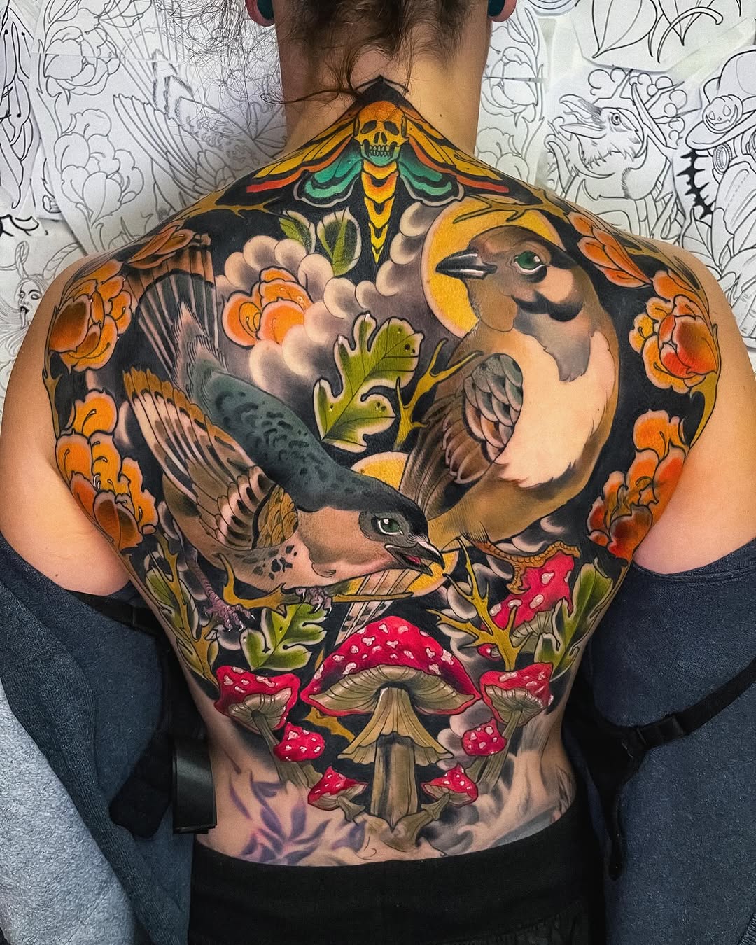 Colorful back tattoo featuring birds, flowers, mushrooms, and a skull surrounded by orange blossoms and green foliage on dark skin.