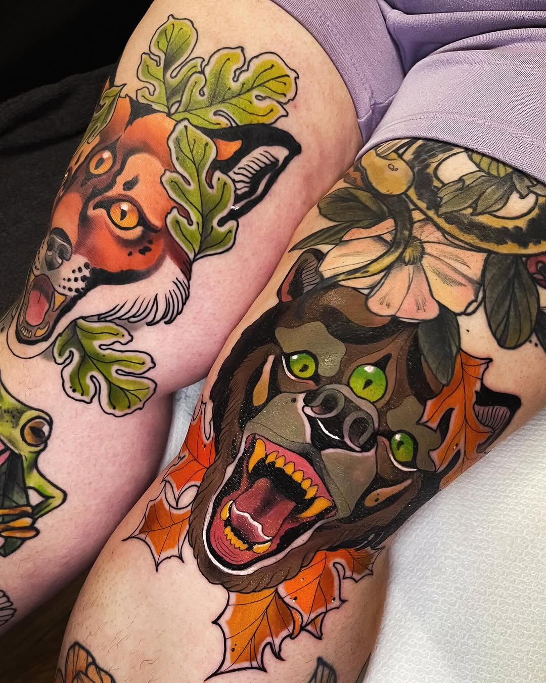 Two vibrant animal head tattoos on thighs featuring an orange cat and brown bear with green eyes, surrounded by autumn leaves and foliage in orange, green, and brown tones.