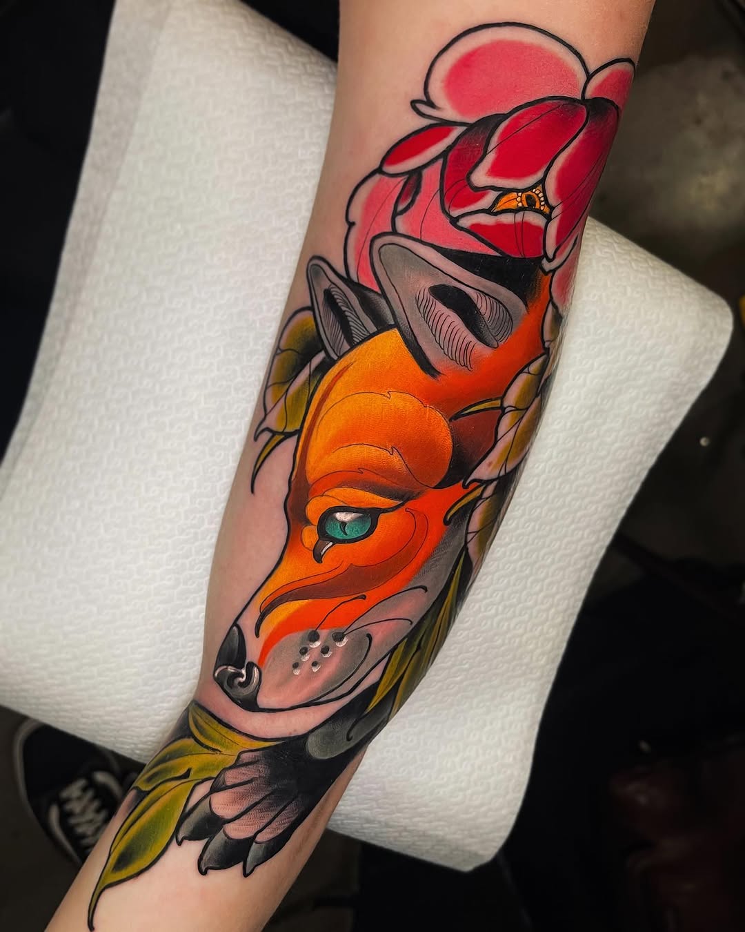 Colorful tattoo on the forearm featuring a layered face design with red petals, orange facial features, and yellow-green leaf elements.