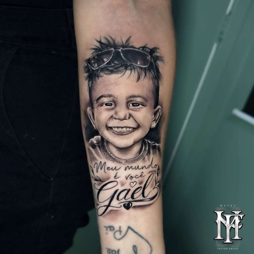 Portrait tattoo of a smiling child wearing oversized goggles on their head, with script text reading "Meu mundo é você Gael" below, placed on the forearm in black ink.
