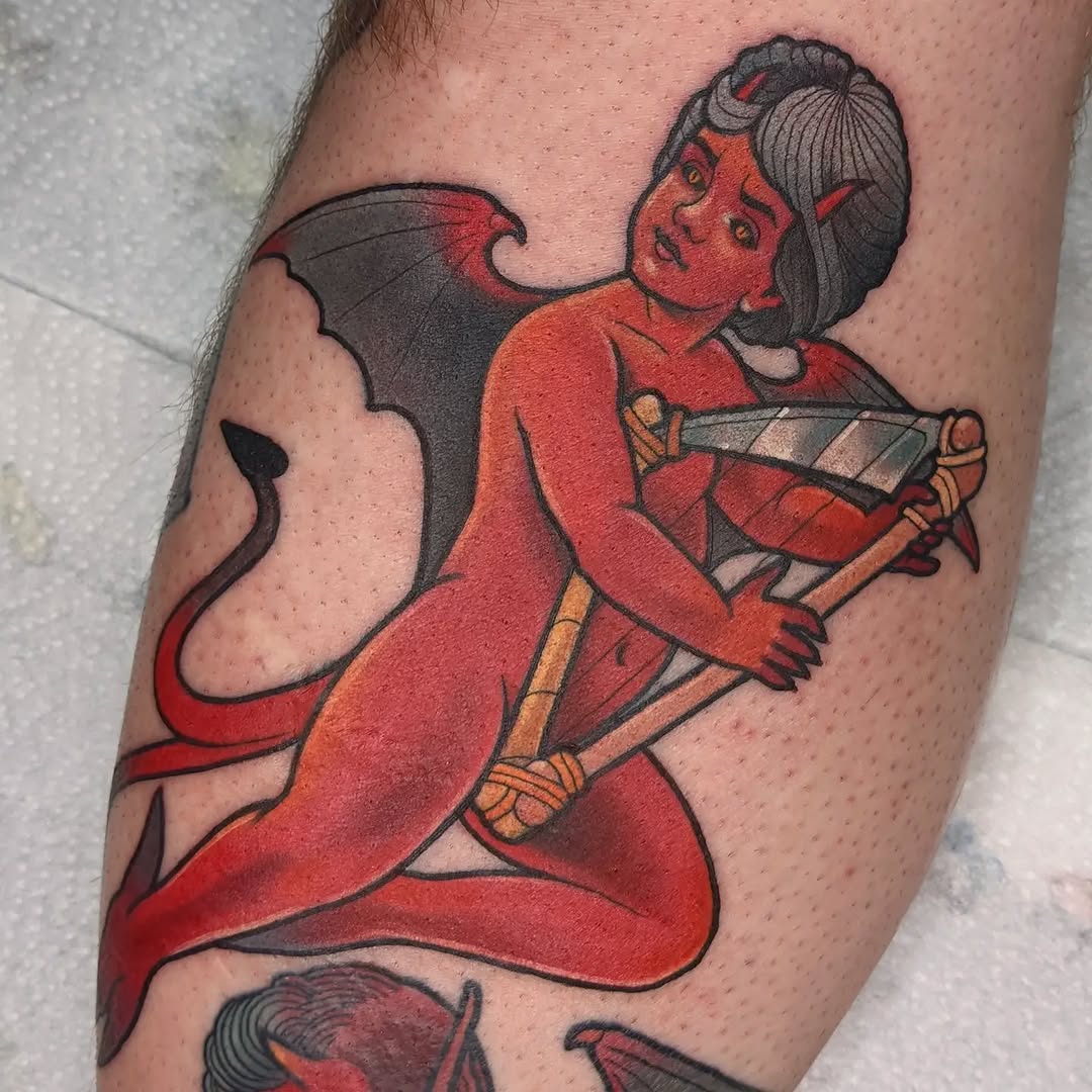 Red-skinned demon woman with black hair and bat wings holding a staff, tattooed on upper leg in red, black, and orange colors.